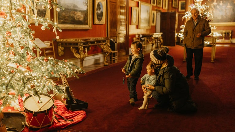 A family walks through a festively room at Attingham Park in Shropshire during Christmas. The scene features twinkling fairy lights strung across trees, historic architecture illuminated in warm tones, and children pointing excitedly at seasonal displays, evoking a joyful holiday atmosphere.